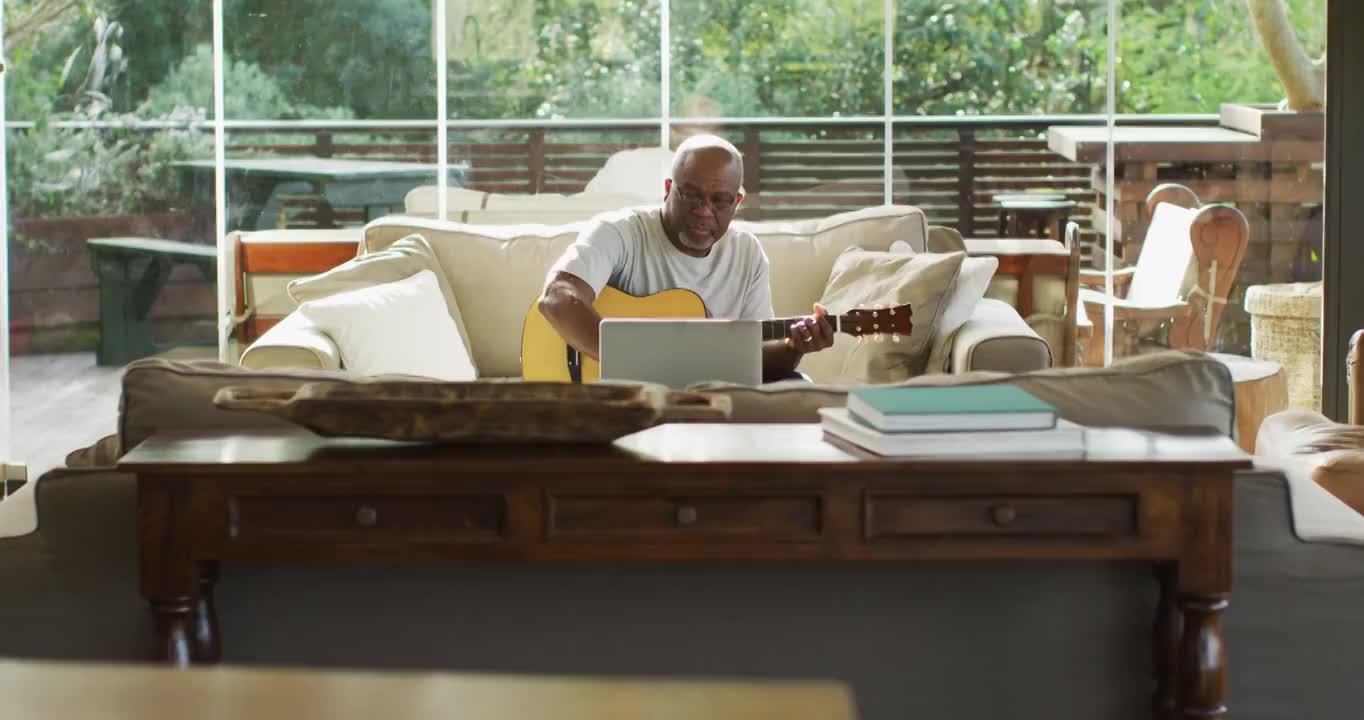 快乐的非洲裔美国老人在客厅玩吉他和使用笔记本电脑视频购买