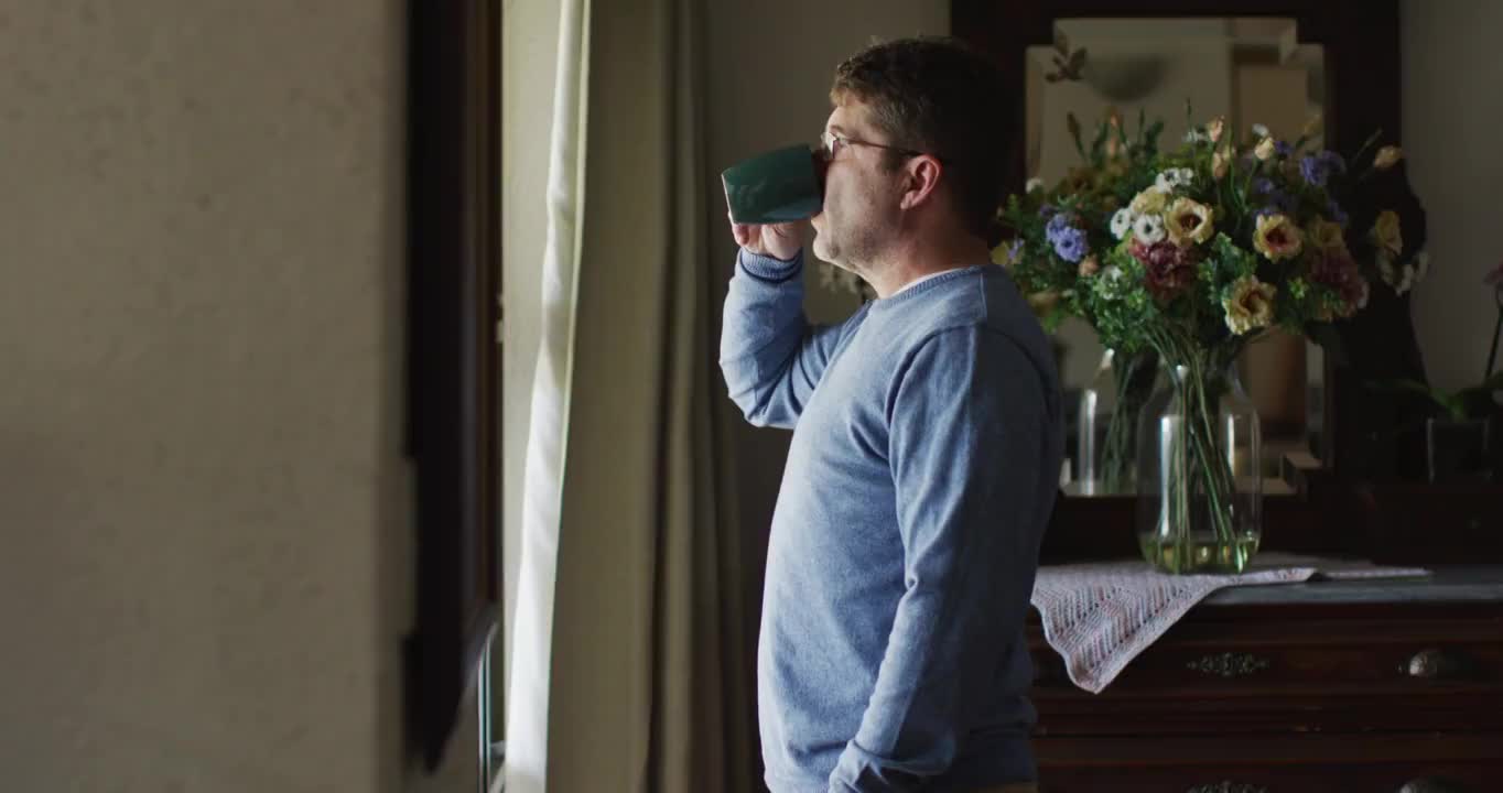 深思熟虑的白人男子站在窗口和喝咖啡在家里视频素材