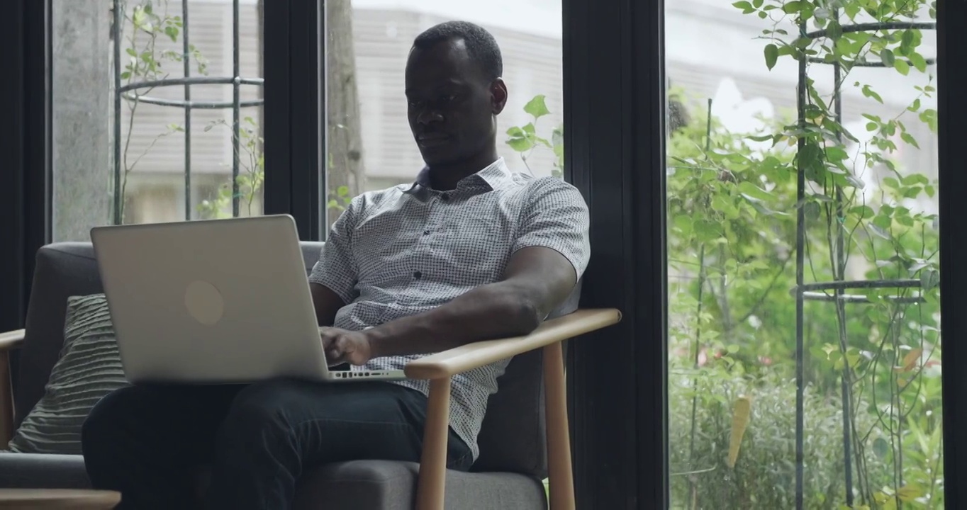 一个黑人坐在靠窗的沙发上，在办公室里使用笔记本电脑。一个非洲视频素材