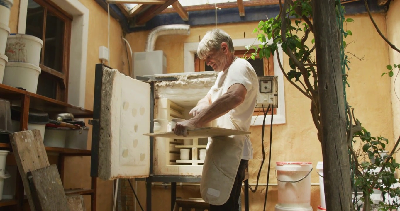 身穿围裙的白人年长男子在陶器作坊从窑中取出烧制的锅视频下载