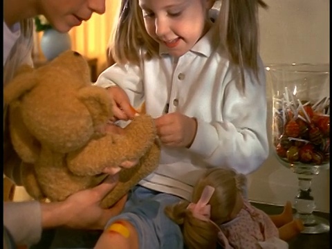 女孩膝盖上缠着胶布，把胶布贴在妈妈抱着的泰迪熊的手上视频素材