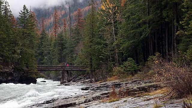 右镜头拍摄的是人们在汹涌的山河上过人行桥，山上有常青树和薄雾。视频素材