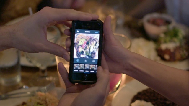 一个年轻人在餐馆里给他的食物拍照，然后用智能手机应用程序编辑视频下载