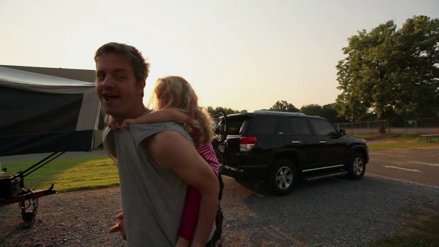 爸爸高兴地旋转着，小女孩躺在他的背上，后面是野营车视频下载