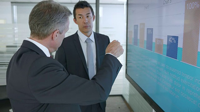 高級白人男性和亞洲男性同事在會議室的大屏幕上討論財務報告中顯示的數字視頻素材