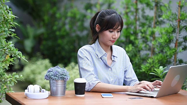 亚洲年轻女子坐在工作场所，在花园里使用笔记本电脑视频素材