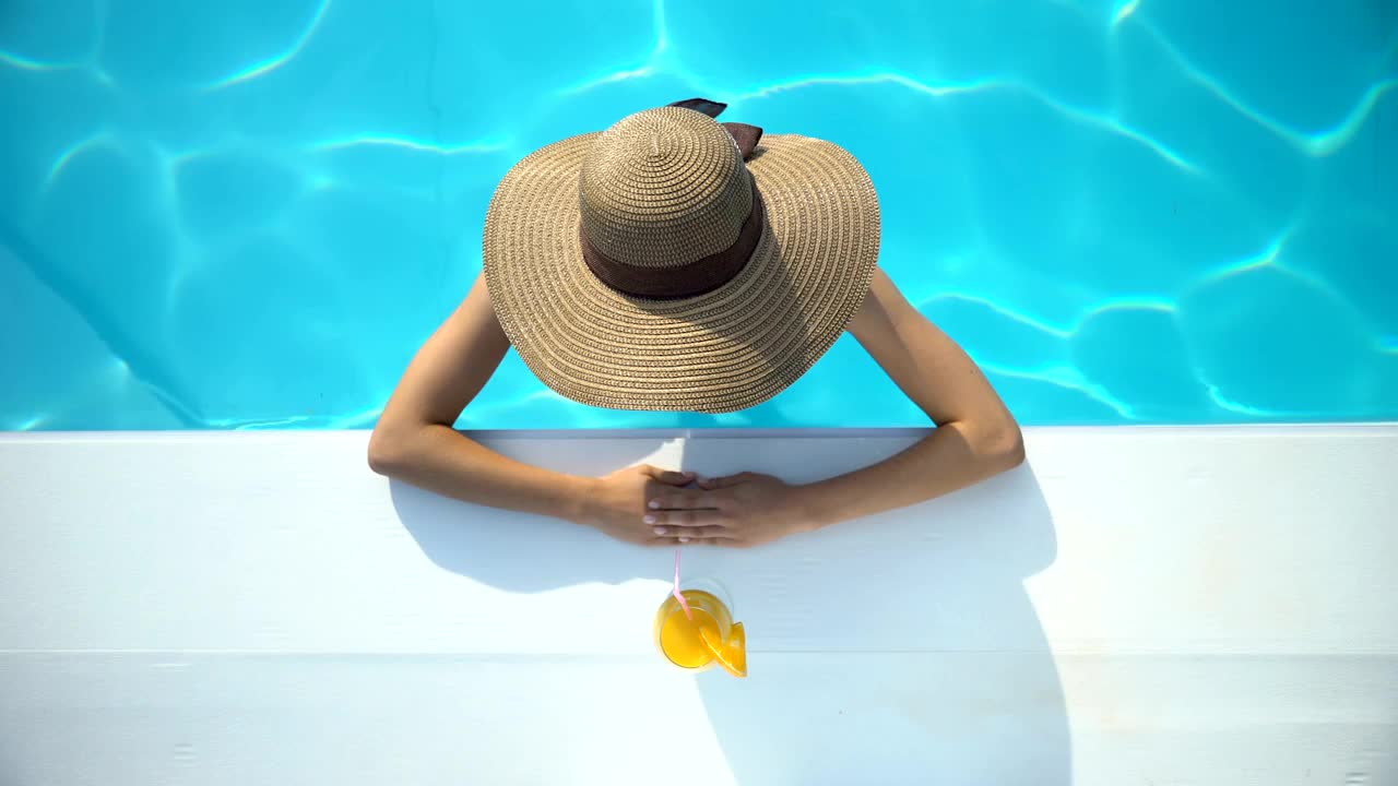 年轻女子戴着优雅的帽子在游泳池游泳，享受美味的鸡尾酒视频下载