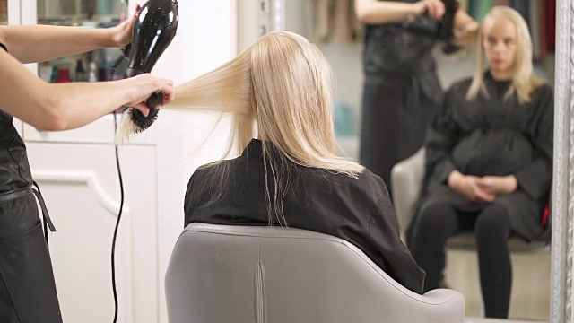 这位怀孕的金发女郎坐在背对着镜头的椅子上。美发师用圆刷把一缕发丝刷成长度，用吹风机吹干。在他们面前的镜子里，倒影是模糊的。视频素材