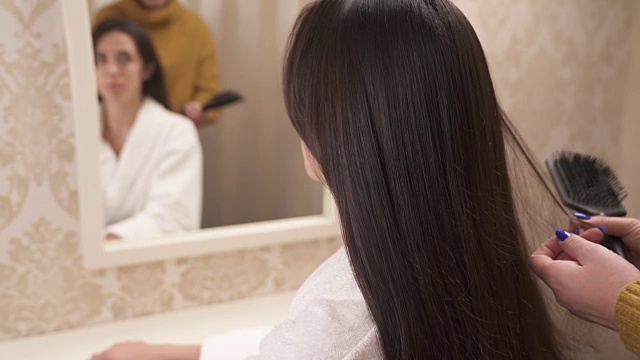 一个迷人的长发黑发女人坐在镜头后面说着什么。一个女人的手把她的头发梳到一定长度，然后解开一根发丝视频素材