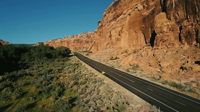无人机跟随银色汽车在高速公路上行驶在陡峭的峡谷岩石山，茂密的沙漠绿化和树木之间。视频素材