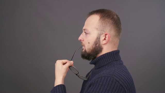 一个年轻的戴着眼镜的白人男子抚摸着他的胡子的特写。人摘下眼镜，把它带到嘴边。孤立在灰色背景。视频素材