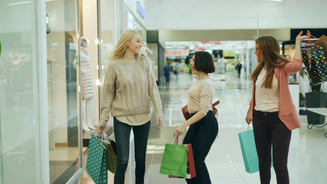 一群快樂的女孩在購物中心玩得很開心，拿著紙袋走著，指著商店櫥窗里的商品笑著跳舞。青春和購物狂理念。視頻下載