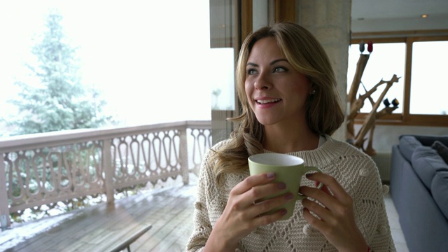 漂亮的年轻女子在家里喝着咖啡，看着冬天的景色，若有所思但微笑着视频素材