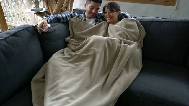一对相爱的年轻夫妇在冬季小屋躺在沙发上，盖着毯子看电视聊天视频下载