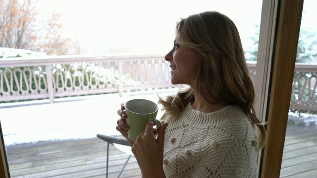 美丽的金发女人望着窗外，一边享受着冬日的景色，一边喝着咖啡视频素材