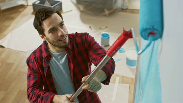 一个穿着棕色牛仔裤和红色格子衬衫的男人正在用滚轴刷墙。油漆颜色为浅蓝色。室内装修。从上面。视频素材