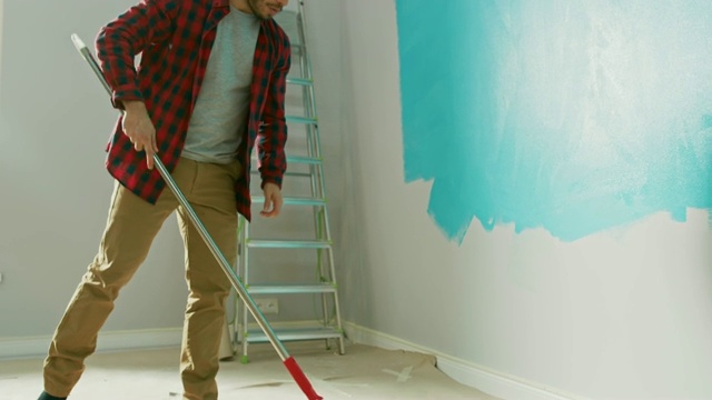 一名穿着棕色牛仔裤和红色格子衬衫的男子在一个长柄滚筒上涂了浅蓝色油漆，开始刷墙。油漆颜色为浅蓝色。他边工作边唱歌。家里装修。视频素材