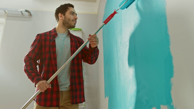 一个穿着棕色牛仔裤和红色格子衬衫的男人正在用滚轴刷墙。油漆颜色为浅蓝色。他边工作边唱歌。室内装修。视频素材