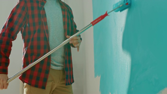 一名穿着棕色牛仔裤和红色格子衬衫的男子在一个长柄滚筒上涂了浅蓝色油漆，开始刷墙。油漆颜色为浅蓝色。室内装修。视频素材