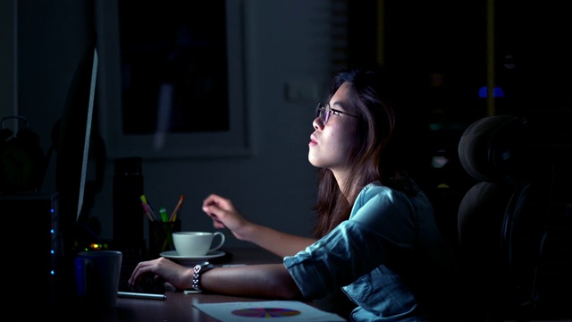 迷人的亚洲女人工作到很晚，喝咖啡与严肃的行动在桌子前的电脑显示器桌面在黑暗的工作场所，工作到很晚和努力的概念视频下载