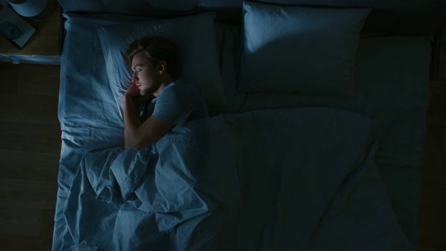 俯视图英俊的年轻人舒适地睡在床上在他的卧室晚上。蓝色的夜间颜色和寒冷微弱的灯柱灯光透过窗户。视频购买