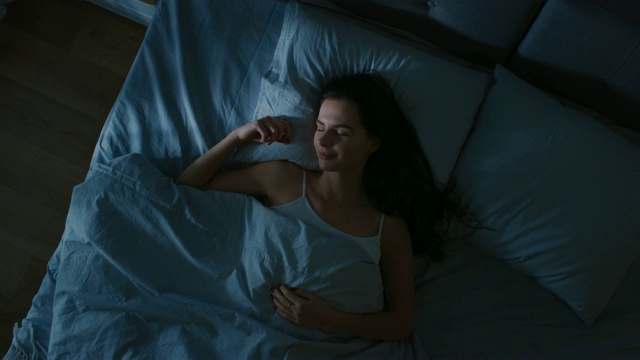 俯视图美丽的年轻女子舒适地睡在床上在他的卧室晚上。蓝色的夜间颜色和寒冷微弱的灯柱灯光透过窗户。视频购买