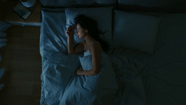 俯视图美丽的年轻女子舒适地睡在床上在他的卧室晚上。蓝色的夜间颜色和寒冷微弱的灯柱灯光透过窗户。视频购买