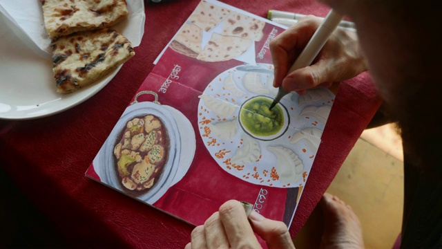 一名男子坐在咖啡馆里，用艺术记号笔在速写本上画出印度食物的图像视频下载