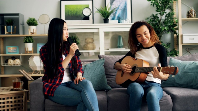 亚裔和非裔美国女性在自由时间一起在家里弹吉他和遥控唱歌。多民族友谊和音乐理念。视频素材