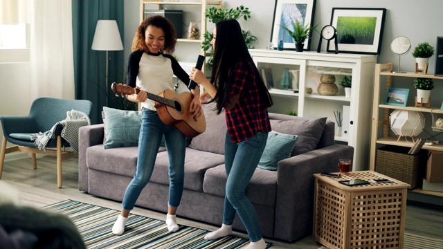 可爱的年轻亚洲和非洲裔美国女士在家里遥控唱歌和弹吉他放松表达积极的情绪。友谊和乐趣的概念。视频素材