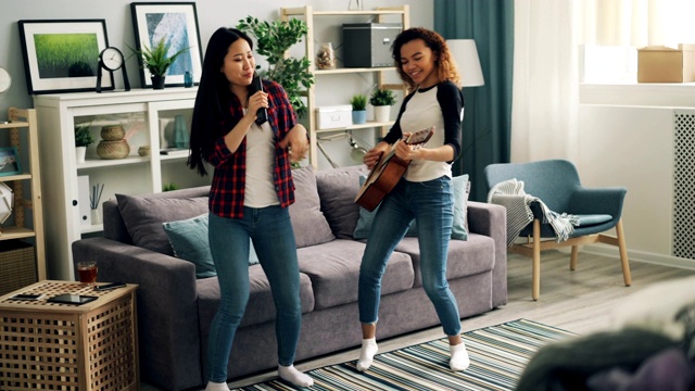 快乐的年轻妇女非洲裔美国人和亚洲人在休闲服装弹吉他，跳舞和唱歌在电视遥控器在现代公寓的乐趣。视频下载