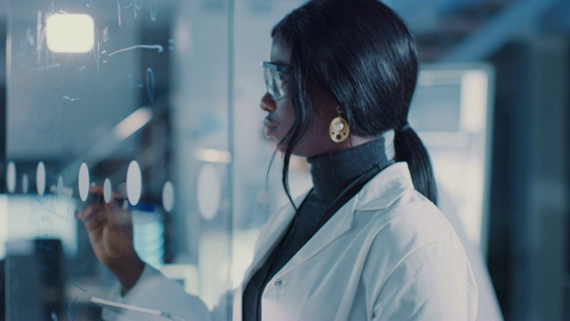 在研究实验室聪明美丽的非裔美国女科学家穿着白大褂和防护眼镜在玻璃白板上写公式，参考她的平板电脑视频下载