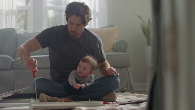 一名男子盘腿坐着，膝盖上放着婴儿，他正用螺丝刀组装平板家具。视频下载