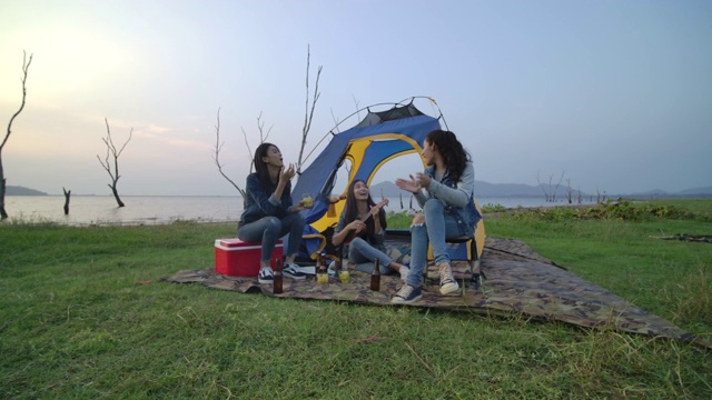 慢镜头——一群年轻的亚洲女孩在湖边露营，在帐篷前弹吉他，一起唱歌。露营，旅行和友谊的概念。视频素材