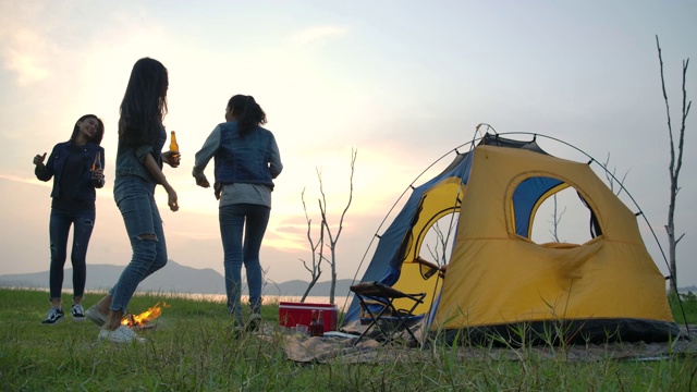 慢动作-一群年轻的亚洲女孩在湖边的帐篷前露营，饮酒和跳舞。露营，旅行和友谊的概念。视频素材