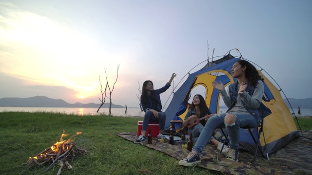 慢镜头——一群年轻的亚洲女孩在湖边露营，在帐篷前弹吉他，一起唱歌。露营，旅行和友谊的概念。视频素材