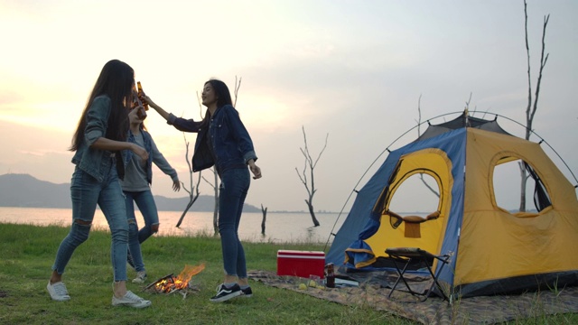 慢动作-一群年轻的亚洲女孩在湖边的帐篷前露营，饮酒和跳舞。露营，旅行和友谊的概念。视频素材