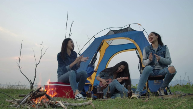 慢镜头——一群年轻的亚洲女孩在湖边露营，在帐篷前弹吉他，一起唱歌。露营，旅行和友谊的概念。视频购买