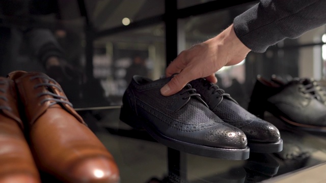 一个男性顾客从奢侈品商业时尚商店的玻璃陈列柜里拿起一双经典风格的深蓝色鞋子的特写。接近相机拍摄视频下载