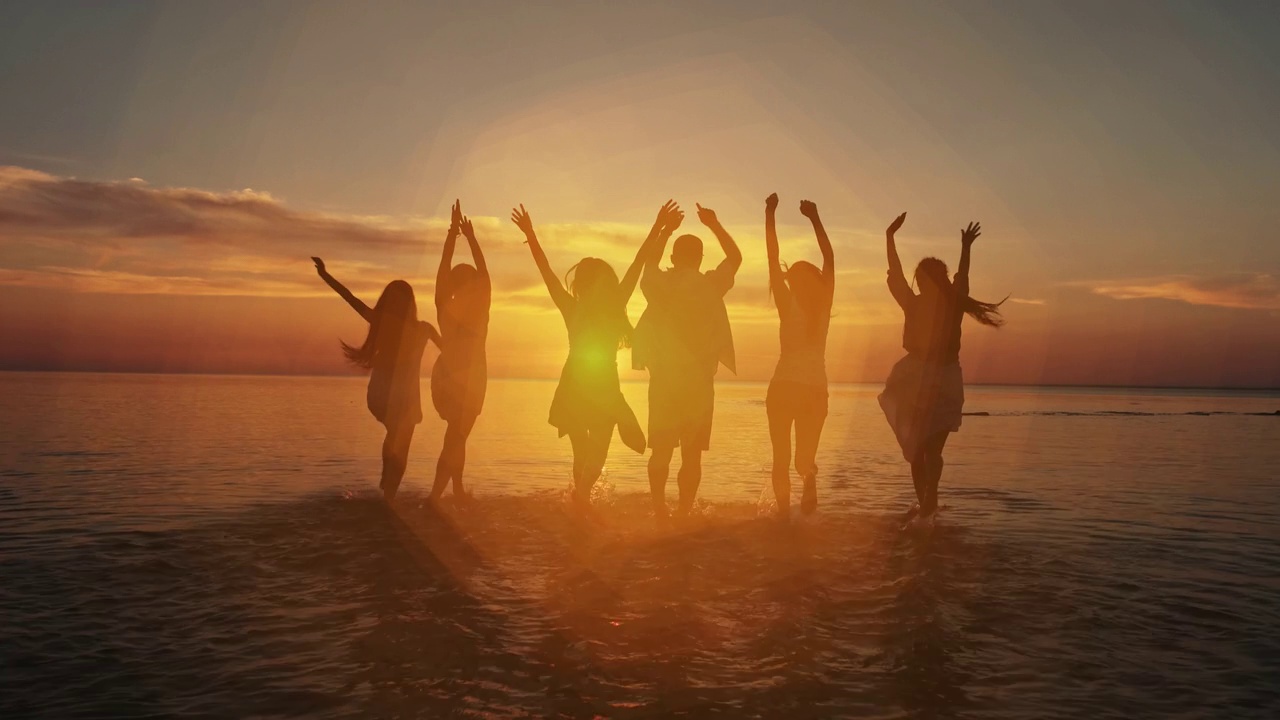 一群快乐的女孩和男人在海边的水里奔跑嬉戏，在夕阳下跳舞，洒落在夏日的夕阳下。海滩派对。太阳耀斑。慢动作视频下载