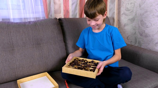 男孩坐在家里的沙发上打开一盒巧克力糖果。视频素材