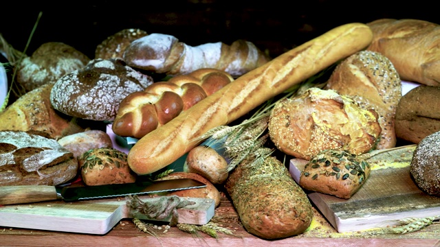 各种各样的新鲜面包与小麦和刀切面包在黑暗的木制背景。在前景的硬壳面包，法棍面包，编织面包和玉米面包。视频素材