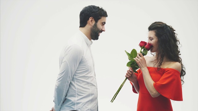 一对美丽浪漫的情侣被孤立在白色的背景上。一个迷人的年轻女子穿着红玫瑰的裙子和一个美丽的男子穿着白衬衫送玫瑰的爱和温柔。情人节!视频素材