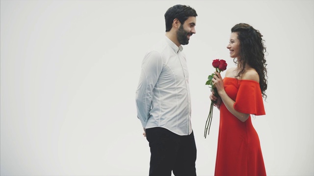 一对美丽浪漫的情侣被孤立在白色的背景上。一个迷人的年轻女子穿着红玫瑰的裙子和一个美丽的男子穿着白衬衫送玫瑰的爱和温柔。情人节!视频素材