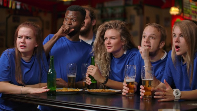 一群穿着蓝色t恤的男女粉丝在酒吧里喝着啤酒，欢呼雀跃。非裔美国人和欧洲人视频素材