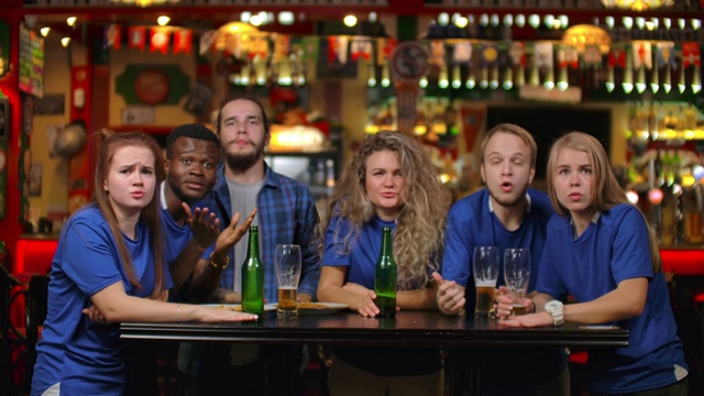 啤酒吧里穿着蓝色衬衫的情绪激动的球迷。一个多种族的非裔美国人群体因为他们团队的失败而感到沮丧和悲伤。击败你最喜欢的足球，排球，视频素材