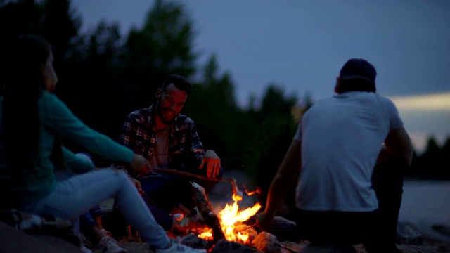 四个年轻的朋友有乐趣烤棉花糖棍棒和交谈坐在篝火在农村一起视频素材