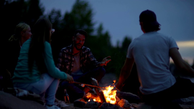 四个年轻的朋友有乐趣烤棉花糖坐在篝火在农村。快乐的男人帮助女性朋友把棍子塞进棉花糖里视频素材