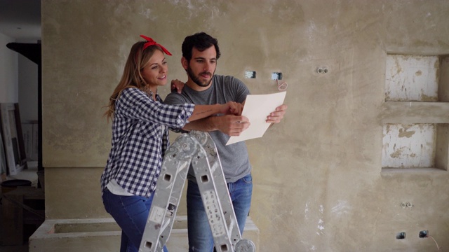 一对快乐的夫妇正在进行家庭装修，一边看着纸上的设计，一边指着一堵墙说话视频下载