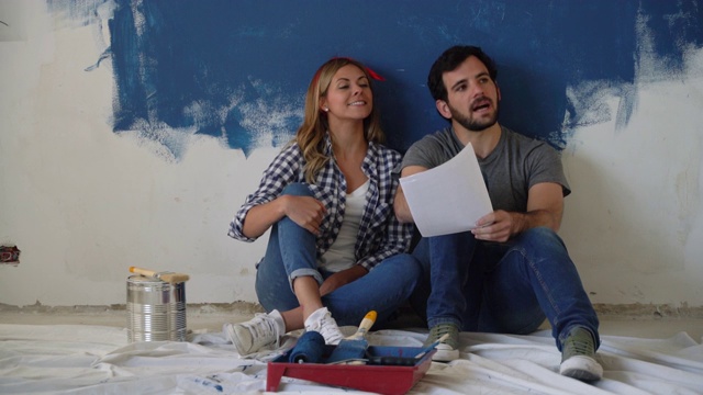 一对幸福的夫妇坐在地板上看着纸上的设计在家庭装修期间视频素材